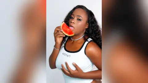 Beautiful woman in a white tank top eating a fresh watermelon slice against a plain white background.