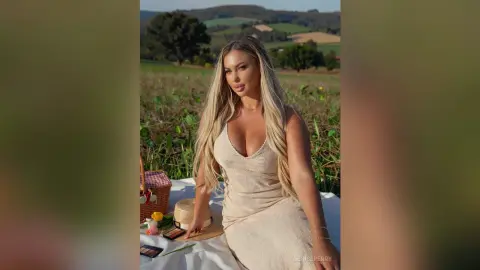 Blonde woman in a beige sleeveless dress relaxing by a picnic basket in a sunny rural field.