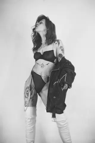AlisaMoss poses in black lingerie and a leather jacket, showing off her tattoos and long hair against a plain background.