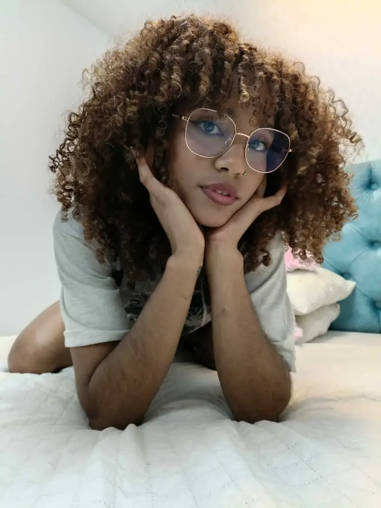 CinthiaWalker poses with her curly dark hair framing her face, showing her bare shoulders and neck against a purple background.