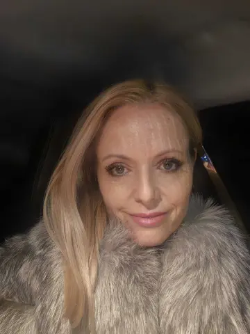Smiling blonde woman in a cozy grey faux fur coat sitting in a car at night.