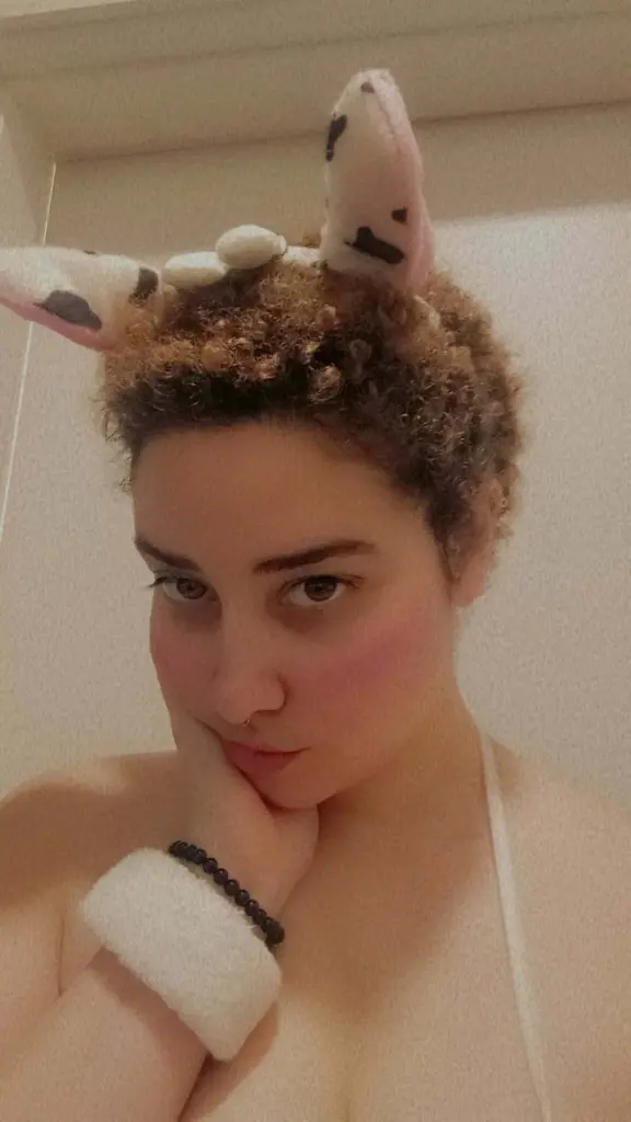 MielMal looks at the camera with a soft expression, showing her face and curly hair against a plain wall.