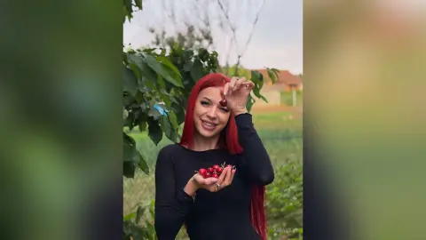 AriaVex smiles while holding red berries in her hands against a background of green leaves and trees.