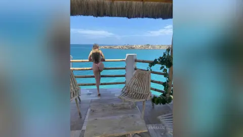 Woman in pink bikini relaxing on a wooden balcony overlooking a tropical turquoise ocean view.