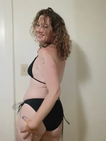 Smiling woman with curly hair wearing a black bikini in a bright indoor setting.