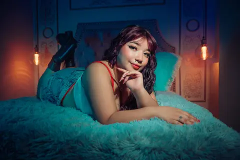 Smiling young woman in denim shorts and white top relaxing on a teal faux fur bed in moody, warm lighting.