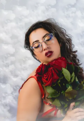 Dreamy portrait of a woman with glasses holding red roses against a soft cloudy sky background.