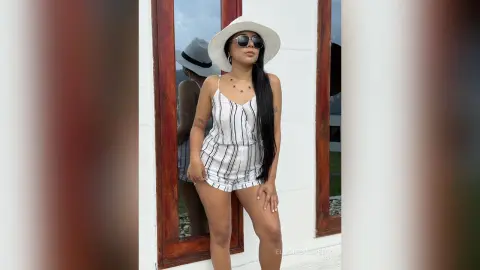 Stylish woman in white striped romper and sun hat posing outdoors in bright summer sunlight.