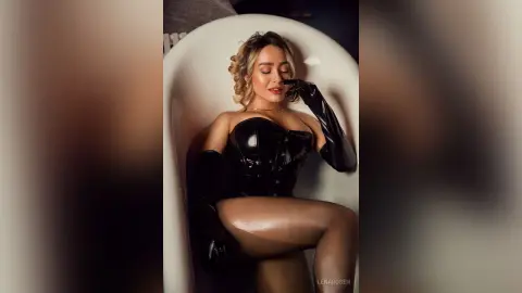Glamorous woman in a black latex corset and long gloves relaxing in a white bathtub with a moody aesthetic.