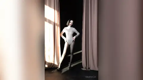 Elegant woman in a white bodysuit and black tights posing in a dramatic, sunlit studio setting.