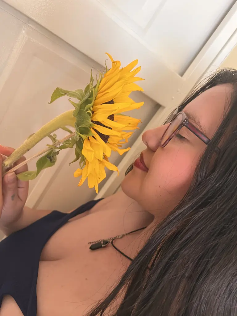 Lullabloom — Lullabloom holds a large yellow sunflower near her face, wearing a dark blue tank top and glasses.