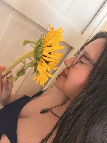 Young woman in a black tank top smelling a bright yellow sunflower in a cozy indoor setting.