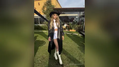 Stylish woman in a black leather trench coat and white outfit posing in a sunny garden setting.