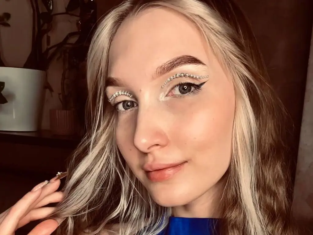 Blonde woman with sparkly rhinestone eye makeup and winged eyeliner posing in a warm indoor setting.