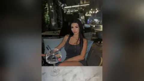 Elegant woman in a dark grey tank top enjoying a cocktail at a dimly lit outdoor restaurant patio.