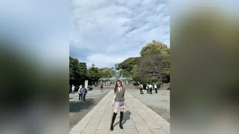 Young woman in a checkered vest and skirt standing before a giant Buddha statue at a serene temple garden.