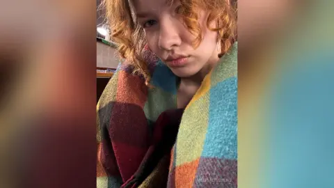 Redheaded woman wrapped in a colorful plaid blanket looking cozy and relaxed indoors.