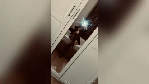 Woman in black outfit sitting on a bed in a modern bedroom, captured in a moody mirror selfie.