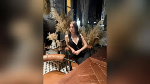 Young woman in a chic black sleeveless dress sitting at a wooden bar in a cozy, modern restaurant setting.