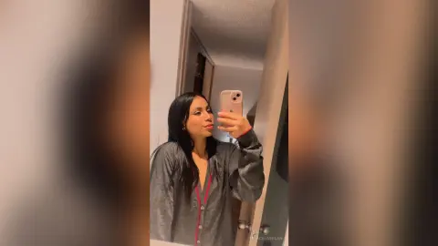 Young woman with long dark hair wearing a grey button-down shirt taking a mirror selfie in a dimly lit room.