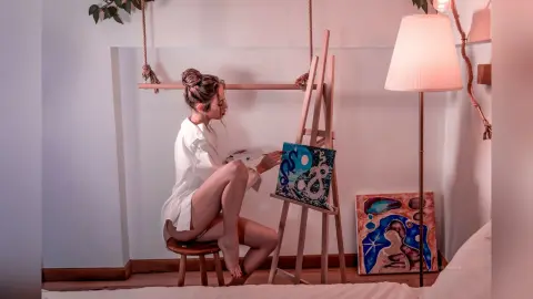 EvelynMills sits on a wooden stool in a white shirt, painting on a canvas set up on an easel.