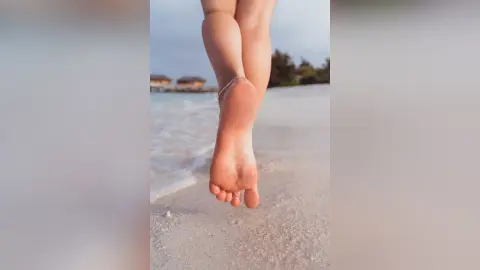 Elie shows off her bare foot and toes while walking along a sandy beach near the ocean water.