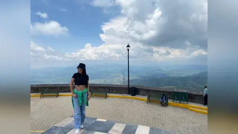 Woman in black crop top and light jeans standing on a scenic mountain lookout under a cloudy blue sky.