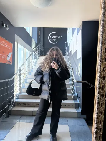 Woman in black puffer jacket and trousers taking a mirror selfie in a modern minimalist staircase lobby.