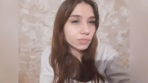 Young woman with long brown hair wearing a cozy white hoodie against a floral patterned wallpaper background.