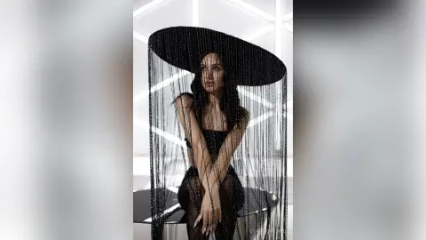 Elegant woman in a black sequined outfit and wide-brimmed fringed hat posing in a minimalist white studio.