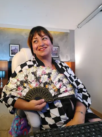 Smiling woman in a black and white checkered kimono holding a decorative floral hand fan at a desk.