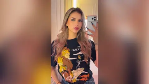 Young woman with long wavy hair wearing a black Lion King graphic t-shirt taking a mirror selfie in a bright room.