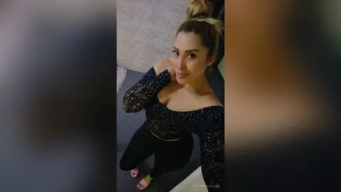 PelinnaCollins poses in a black sparkly long-sleeve crop top and black leggings, looking up at the camera with her hair in a bun.