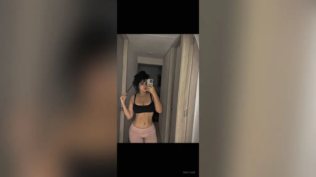 EmilyJune — EmilyJune poses in a black sports bra and pink leggings, taking a mirror selfie in a hallway.