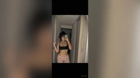 EmilyJune poses in a black sports bra and pink leggings, taking a mirror selfie in a hallway.