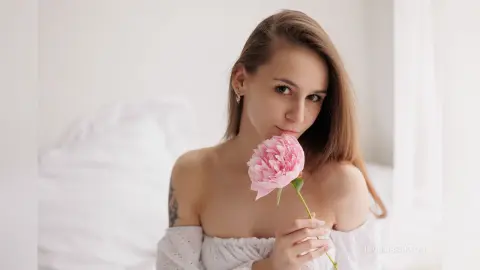 IngridBeckton poses with a pink flower held near her mouth, showing her bare shoulders and long brown hair against a white bed background.