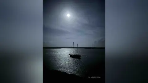 Silhouette of a sailboat on calm ocean waters under a bright full moon in a dark, moody night sky.