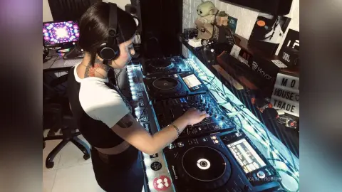 Female DJ wearing headphones and a white crop top mixing music at a professional DJ controller setup.