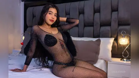 Young woman in a black sheer mesh bodysuit relaxing on a bed in a cozy bedroom setting.