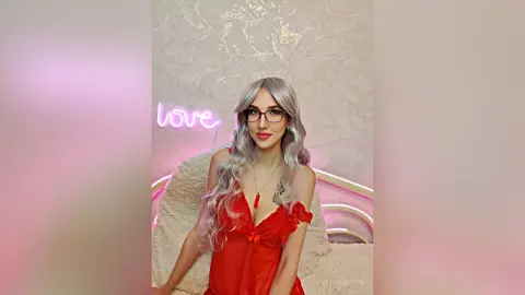 Young woman with long silver hair in a red ruffled dress posing in a dreamy pink room with a neon love sign.