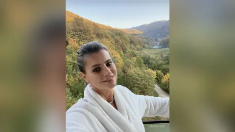 Smiling woman in a white bathrobe relaxing against a scenic autumn forest mountain landscape.