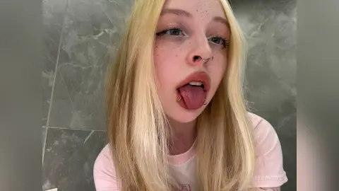 Blonde woman with nose ring sticking out her tongue in a casual pink t-shirt against a grey marble wall.