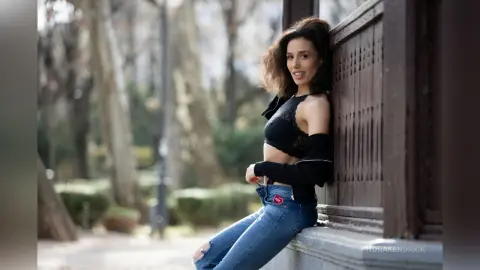 Smiling woman in a black crop top and blue jeans leaning against a wooden fence in a sunny park.
