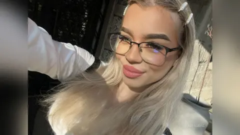 Smiling blonde woman with glasses and pearl hair clips wearing a white top in bright sunlight.