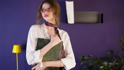 Young woman in white shirt and striped tie holding a book against a purple wall in a moody indoor setting.