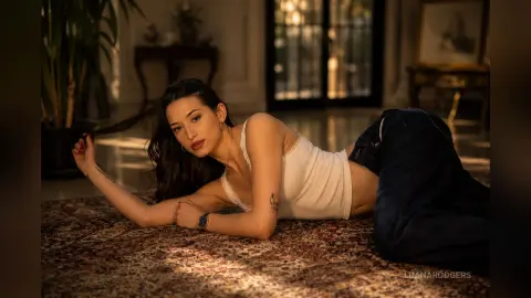 LuanaRodgers lies on a patterned rug in a white tank top and dark jeans, running her hand through her long dark hair.
