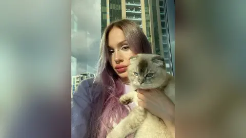 Young woman with long purple hair holding a grey cat in front of a city skyscraper window.