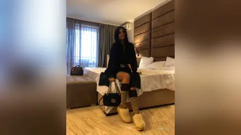 Smiling woman in a black outfit and designer accessories sitting on a bed in a modern hotel room.