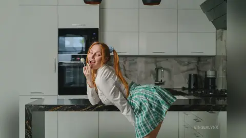 Cheerful redhead woman in a white blouse and green plaid skirt eating a snack in a modern white kitchen.