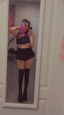 Young woman with pink hair in a black crop top and mini skirt posing in a mirror for a moody selfie.